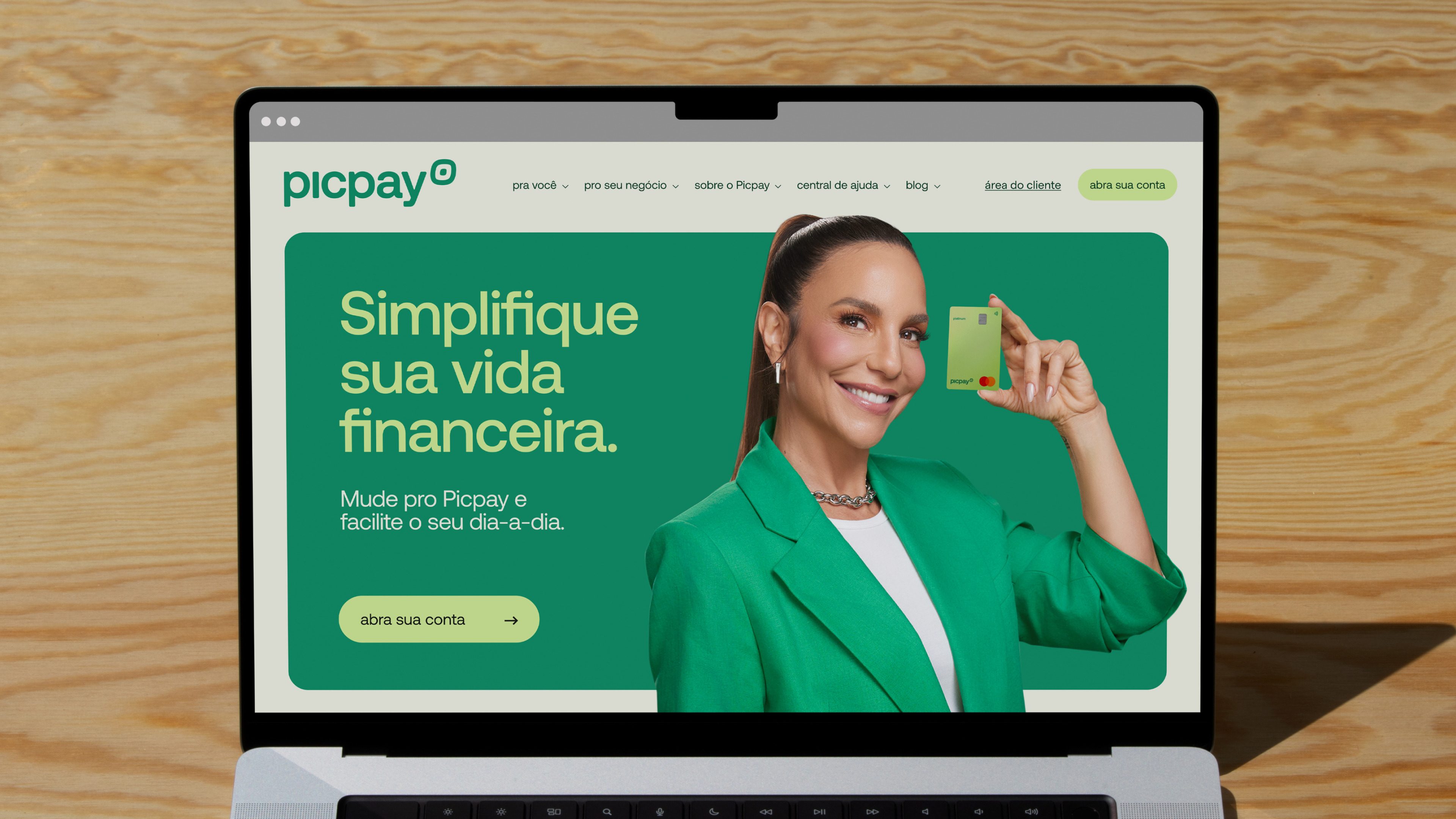 Notebook sobre mesa de madeira com o site do Picpay e uma mulher segurando o cartão verde