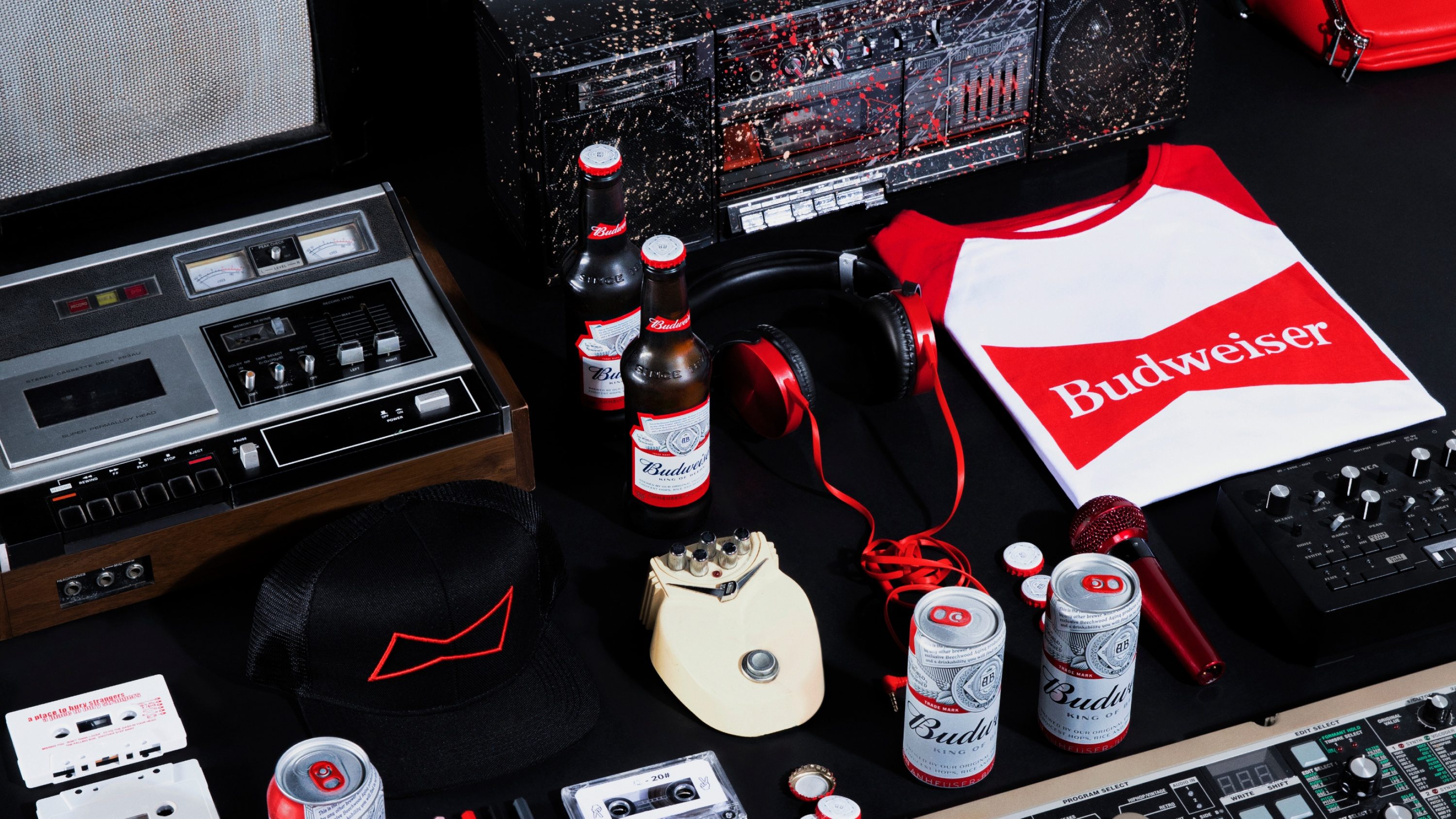Mesa preta com toca-fitas, boombox, fones vermelhos, camiseta, boné, garrafas e latas de Budweiser