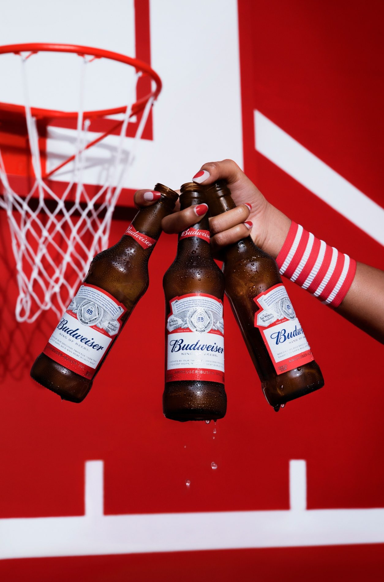 Mão brindando com três garrafas de Budweiser diante de uma cesta de basquete vermelha