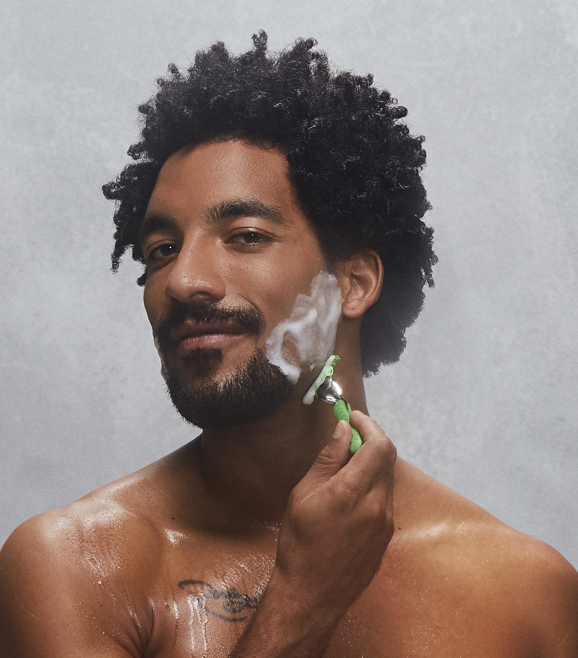 Homem de barba fazendo a barba com um aparelho verde, diante de um fundo cinza