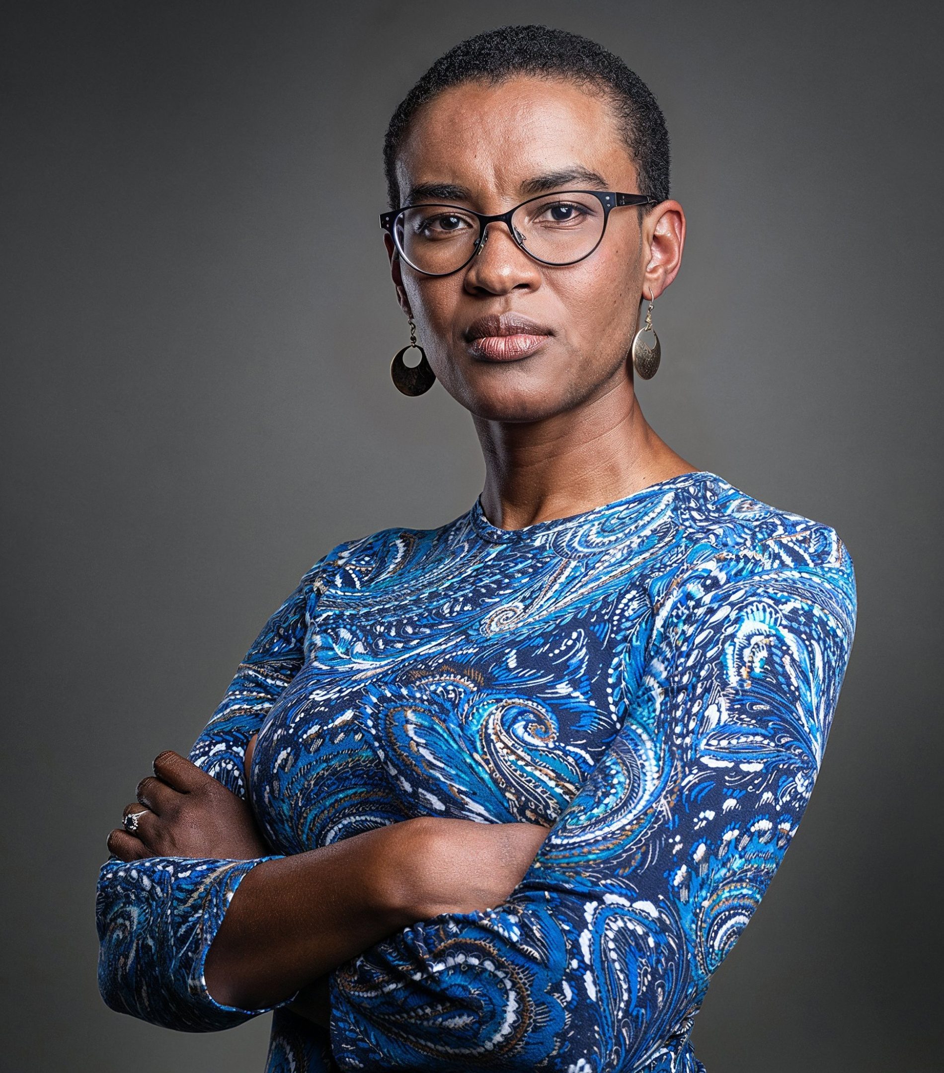 Retrato de uma mulher negra de óculos e braços cruzados, com vestido azul estampado, sobre fundo cinza