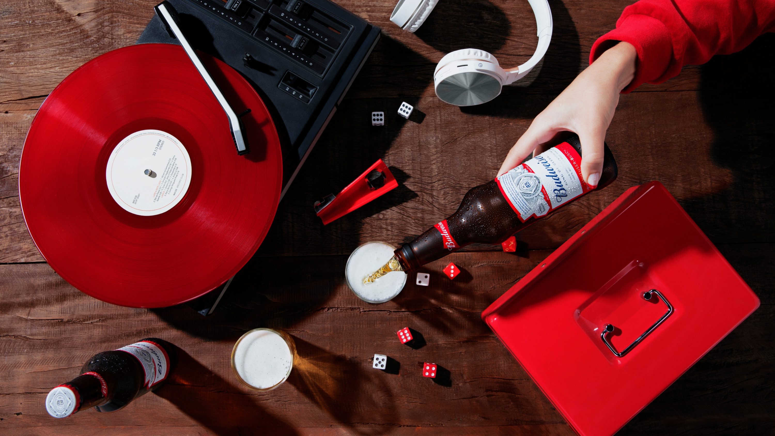 Vista de cima de um toca-discos com vinil vermelho, dados e uma mão servindo Budweiser num copo
