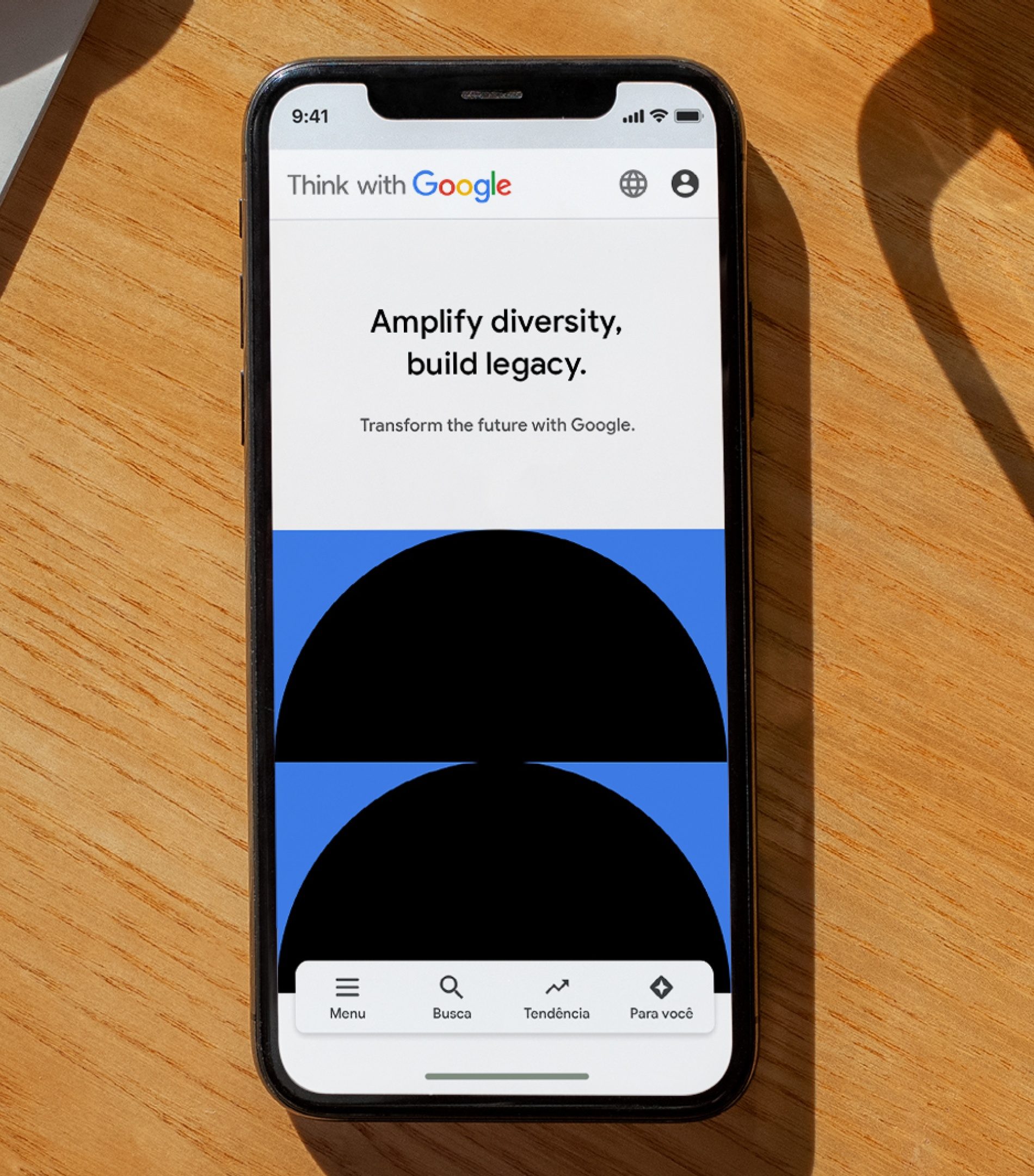 Celular sobre mesa de madeira com o site Think with Google: Amplify diversity, build legacy. e duas meias-luas pretas sobre azul